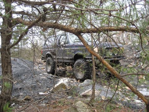 1991 Ford Bronco - A lifted Ford Bronco navigates a rough, off-road path through a wooded area.