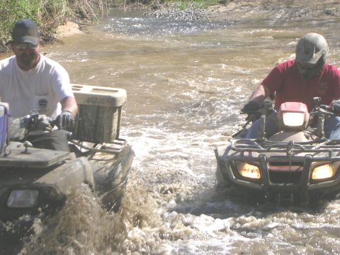 2008 Bombardier Outlander - Two ATVs navigate a muddy stream, kicking up water as they go.
