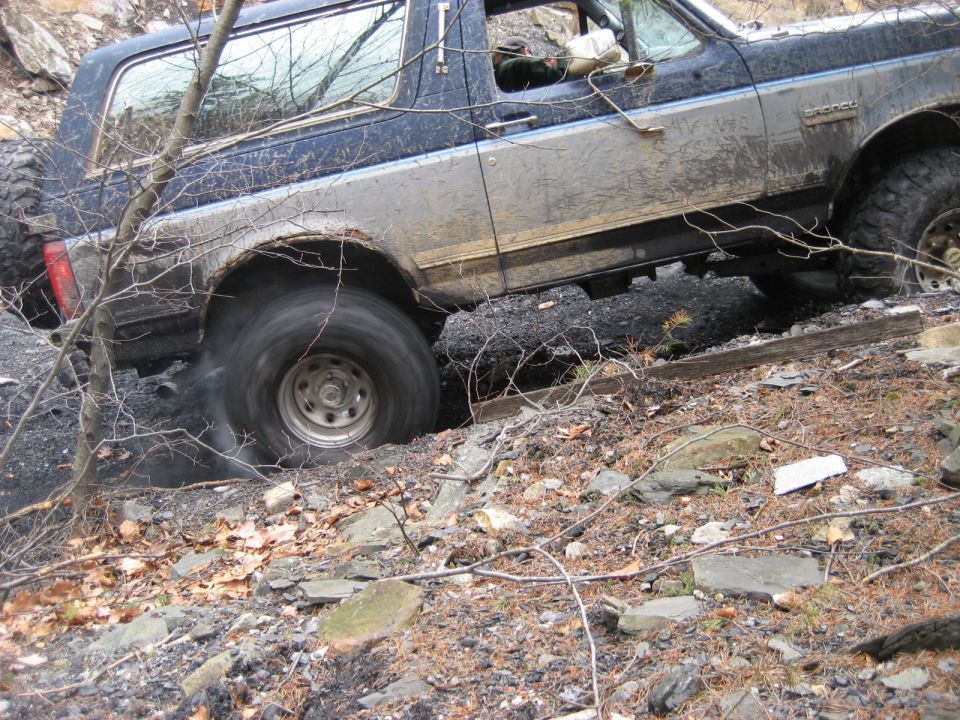 Mud-splattered dark blue and silver Ford Bronco on rocky terrain, driver's side view