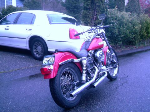1972 Harley-Davidson Super Glide - A pink motorcycle is parked behind a white sedan on a wet street.