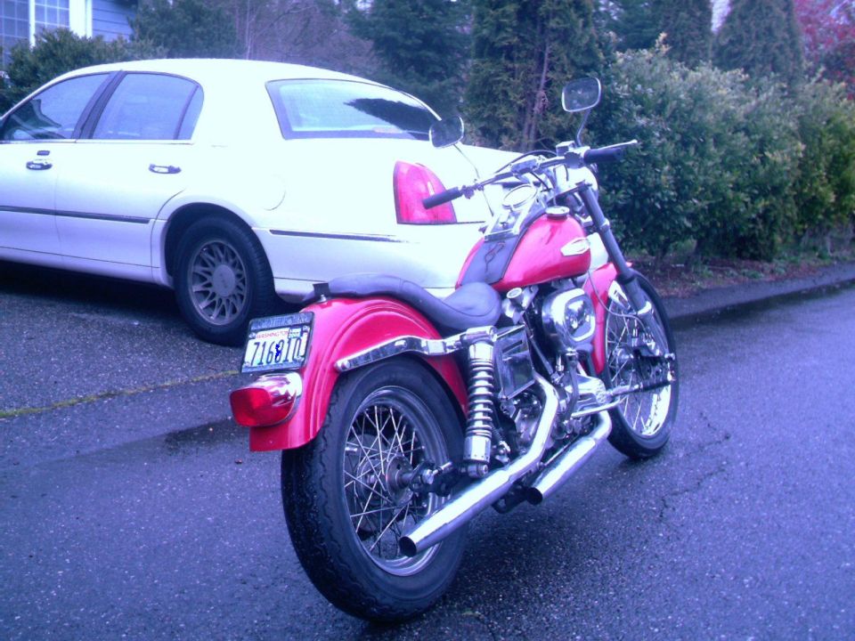 Red Harley motorcycle parked rear view beside white sedan on wet street