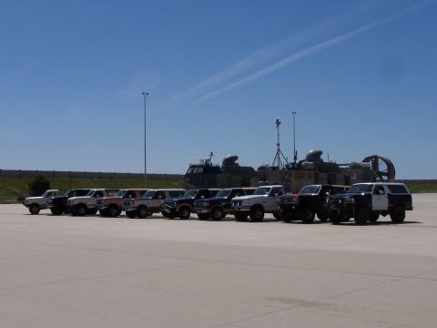 1990 Ford Bronco - Nine full-size Ford Bronco SUVs in white, tan, blue and black lined up diagonally on a wide concrete apron under a clear blue sky. A Navy L…