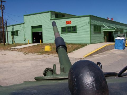 1990 Ford Bronco - A military vehicle's cannon is in the foreground, pointing towards a building with a portable toilet nearby.