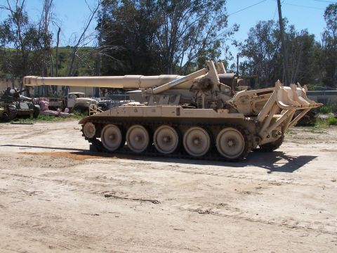 1990 Ford Bronco - A self-propelled howitzer sits in a lot with other vehicles under a clear sky.