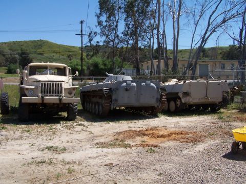 1990 Ford Bronco - Outdoor scene with a tan truck and two grey tracked military vehicles.