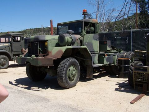 1990 Ford Bronco - Three-quarter front view of a camouflage-painted M-series 6x6 military tractor truck marked "USMC" on the hood, parked on a dirt lot beside…