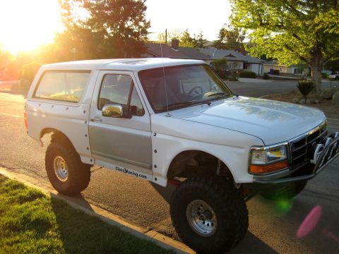 1996 Ford Bronco - White Ford Bronco lifted on oversized mud-terrain tires and polished beadlock-style wheels, parked at a suburban curb at sunset and shot fr…