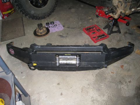 1996 Ford Bronco - A black steel aftermarket winch bumper lying face-up on a garage floor, fitted with a polished roller fairlead and a yellow-tipped D-ring/p…