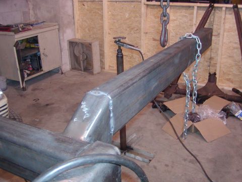 1991 Ford Bronco - A bare truck frame rail sits on jack stands and a hydraulic jack in a wood-framed shop, with a fresh weld bead and grind marks on a boxed r…
