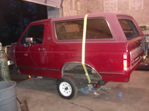 1991 Ford Bronco - This red SUV body is missing its front end and rear axle, and is held up by a yellow strap.