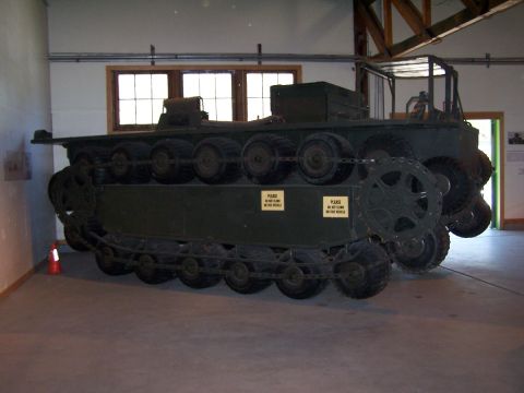 1990 Ford Bronco - Side view of an olive-drab tracked military vehicle displayed indoors on a concrete museum floor, riding on rubber-tired bogie wheels with…