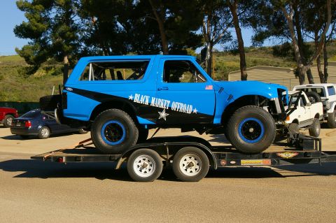 1986 Ford Bronco - A modified blue Bronco is loaded onto a trailer, with other vehicles visible in the background.