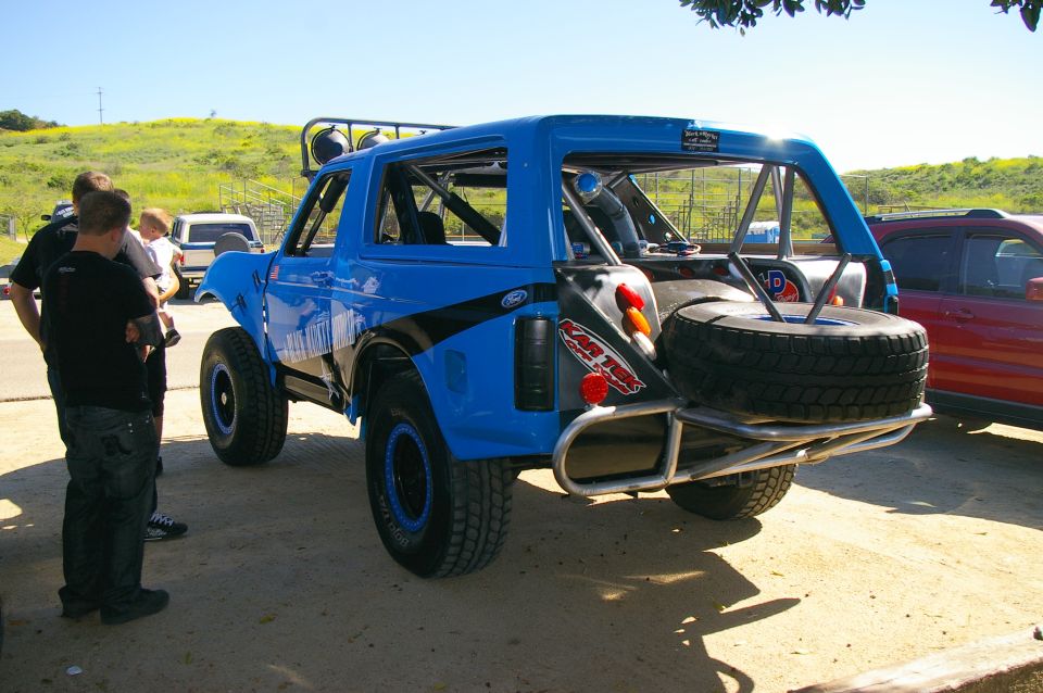 Blue off-road Bronco with roll cage, spare tire on back, and black stripe graphics. People standing nearby