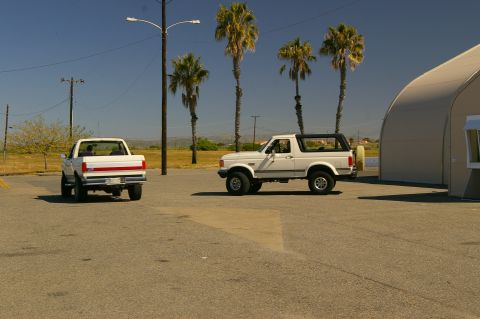 1986 Ford Bronco - A white Ford Bronco and a white Ford pickup truck are parked in a lot with palm trees and a large building in the background.