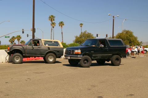 1986 Ford Bronco - Two Ford Broncos are parked on the street with palm trees and traffic lights visible.