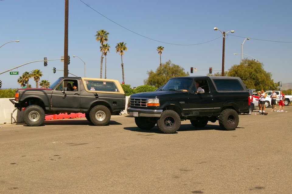 Two Ford Broncos parked on a street, one dark gray with tan top, one black. Palm trees and traffic lights in background