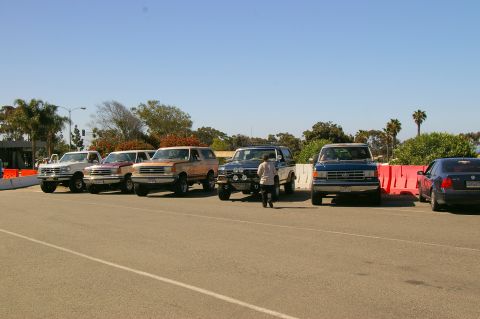1986 Ford Bronco - Several Ford Bronco SUVs and trucks are parked in a lot under a clear blue sky.