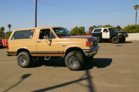 1986 Ford Bronco - Two Ford Broncos parked outdoors on a sunny day, one tan and one two-tone black and white.