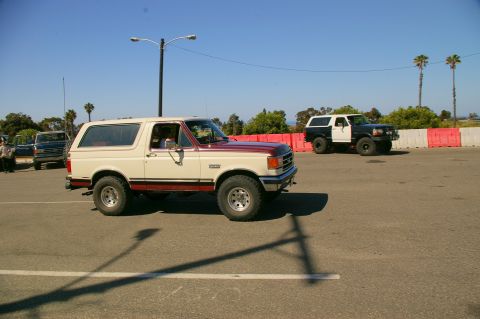 1986 Ford Bronco - Two Ford Broncos are seen driving in a parking lot on a sunny day with palm trees in the background.