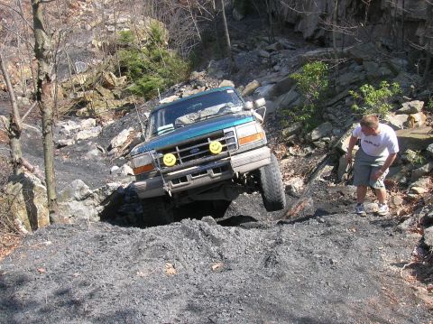1996 Ford Bronco - A teal Ford Bronco is shown off-roading on a steep, rocky trail with a person observing.