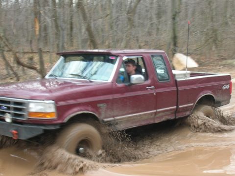 1996 Ford Bronco - A maroon 1996 Ford Bronco navigates a muddy trail, kicking up water and dirt.