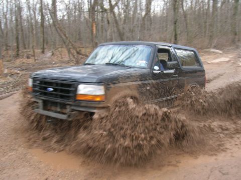 1996 Ford Bronco - A Ford Bronco navigates a muddy off-road trail, sending up a large spray of dirt and water.