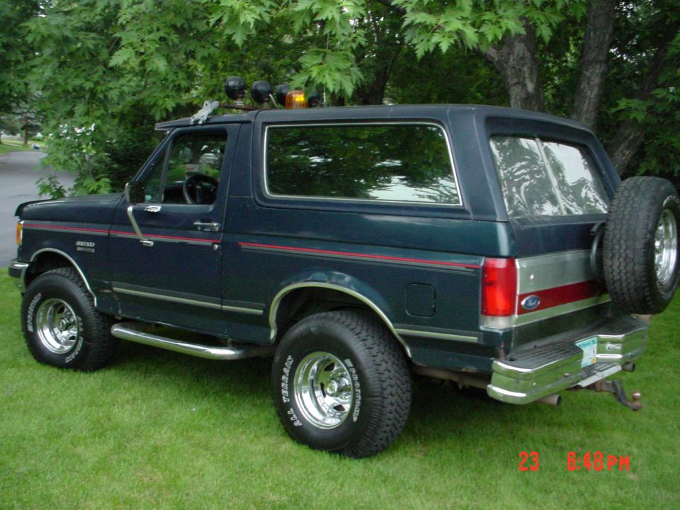 Dark green Ford Bronco SUV on grass, rear three-quarter view with spare tire, roof lights and chrome wheels
