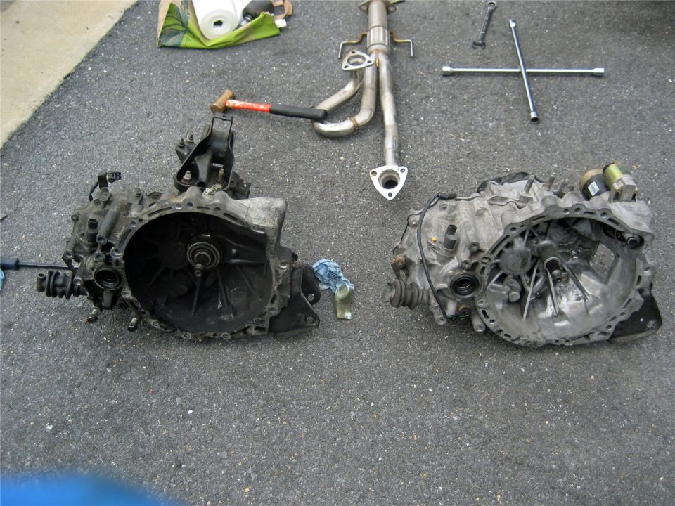 Two car transmissions and an exhaust manifold laid out on asphalt, with a hammer and lug wrench nearby