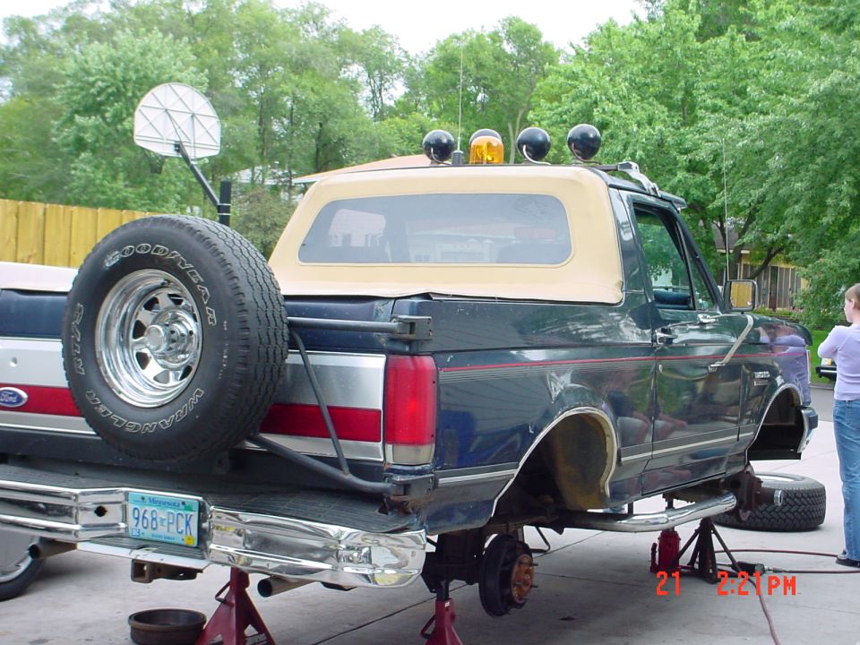 Dark blue Ford Bronco with tan top on jack stands, rear wheels removed, spare tire on swing-away carrier