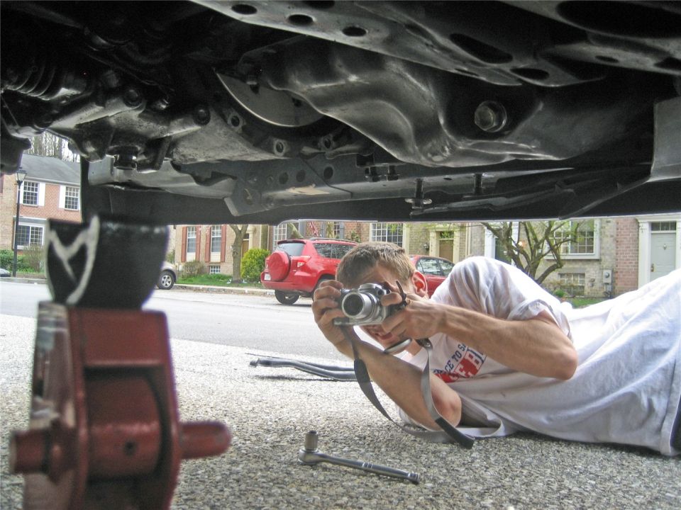 Man lying on ground taking photo of car's undercarriage; red SUV visible in background