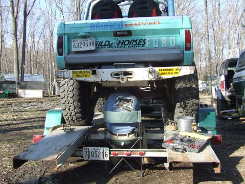1969 Ford Bronco - A person works on a modified Ford Bronco loaded onto a trailer, with tools laid out and another vehicle visible in the background.