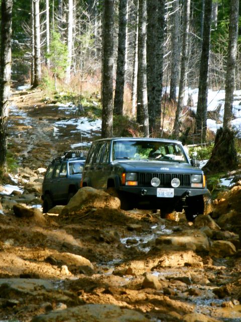 2005 Jeep Wrangler - Two Jeep Cherokee XJs navigate a challenging muddy and rocky off-road path through a forest.