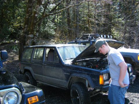 2005 Jeep Wrangler - A mud-streaked dark blue Jeep XJ Cherokee sits with its hood raised on a forest trail, engine bay and inline-six accessories exposed while…