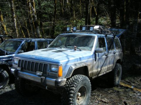 2005 Jeep Wrangler - Three-quarter front view of a lifted two-tone blue Jeep XJ Cherokee Laredo parked on a muddy forest trail, splattered with dried mud. It ca…