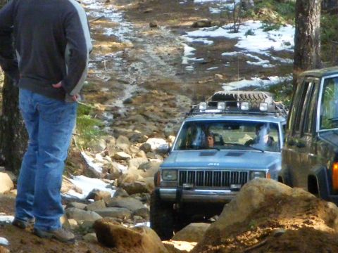 2005 Jeep Wrangler - A blue Jeep XJ Cherokee with a roof rack carrying four round driving lights and a row of recovery gear picks its way up a rocky, partially…