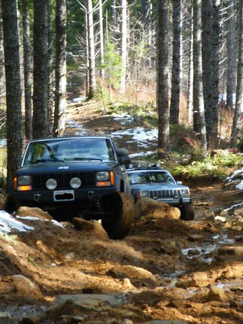 2005 Jeep Wrangler - Two Jeep Cherokees tackle a challenging muddy trail in a forest setting.