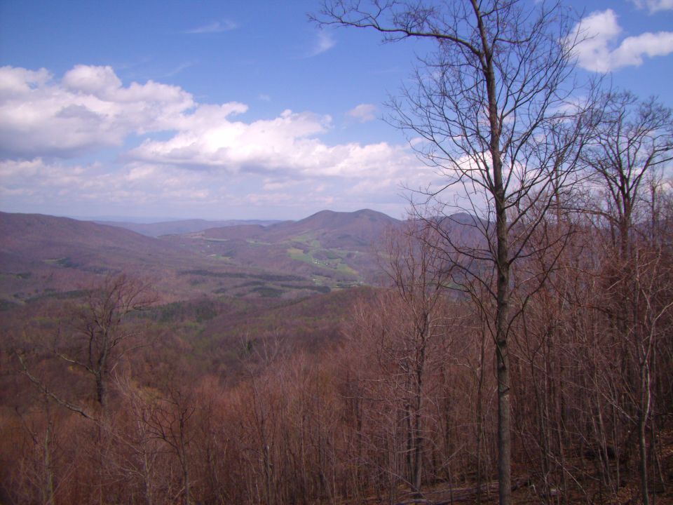 Mountain landscape with bare trees in the foreground and a valley with green fields in the distance under a cloudy blue sky