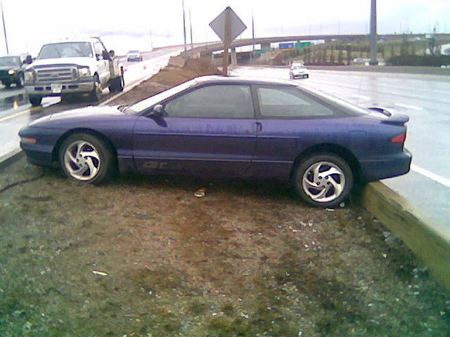 Side view of a 1996 Ford Probe parked on a dirt shoulder next to a highway. A white pickup truck is visible in the background