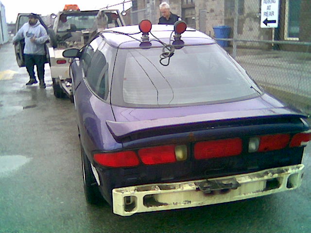 1996 Ford Probe being towed by a flatbed truck, rear view. Tow truck driver stands by the car door