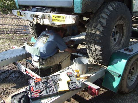 1969 Ford Bronco - A person is working on the undercarriage of a vehicle that is loaded onto a trailer, with tools and a beverage within reach.