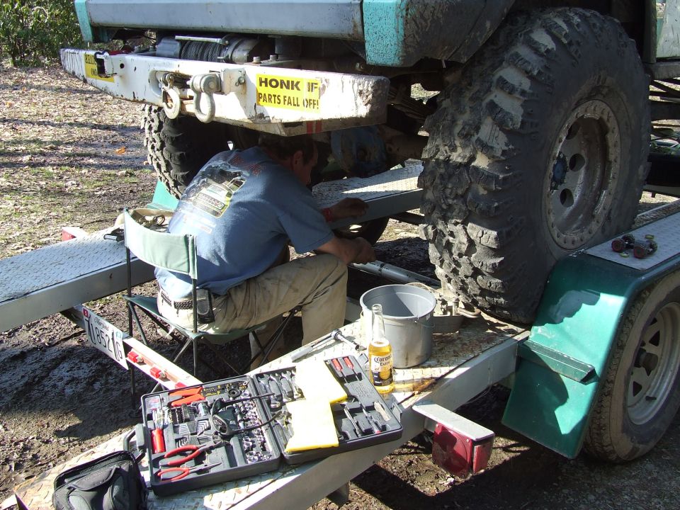 Guy working on the rear axle of a lifted off-road vehicle on a trailer. Tools and a beer are nearby
