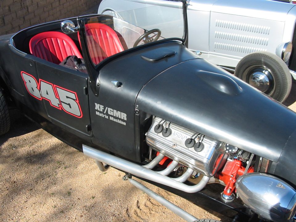 Flat black hot rod roadster #845 with red seats and exposed polished engine, silver vintage car behind