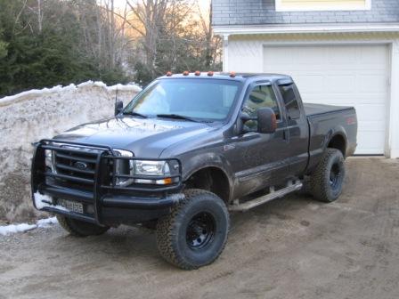 1993 Ford Bronco - Three-quarter front view of a lifted gray Ford Super Duty extended-cab short-bed pickup parked on a snowy gravel driveway beside a white ga…