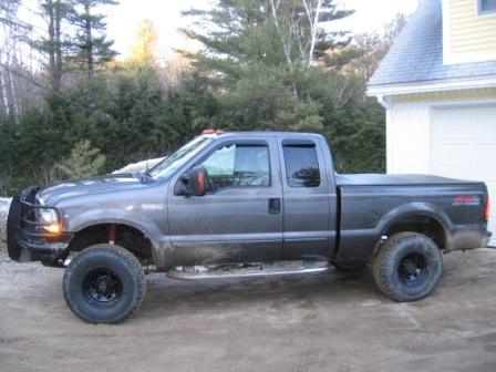 1993 Ford Bronco - Driver-side profile of a lifted gray Ford Super Duty extended-cab short-bed pickup on a dirt driveway beside a yellow house, with pines and…