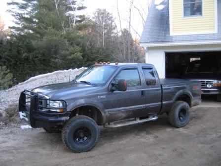 1993 Ford Bronco - Three-quarter front view of a lifted gray Ford Super Duty extended-cab pickup on a dirt driveway in front of an open garage on a yellow hou…