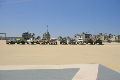 1986 Ford Bronco - Multiple SUVs and pickup trucks are lined up on a sunny day, with large hovercraft visible in the distance.