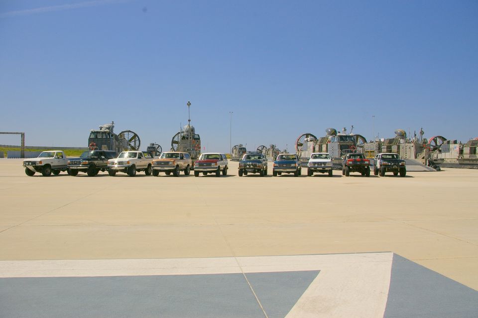 Line of SUVs and pickup trucks, mostly white and tan, parked on a paved lot with hovercraft in the background