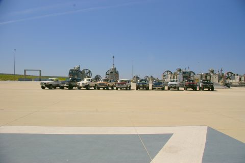 1986 Ford Bronco - A row of various SUVs and pickup trucks are parked on a paved surface, with large hovercraft vehicles visible in the distance under a clear…