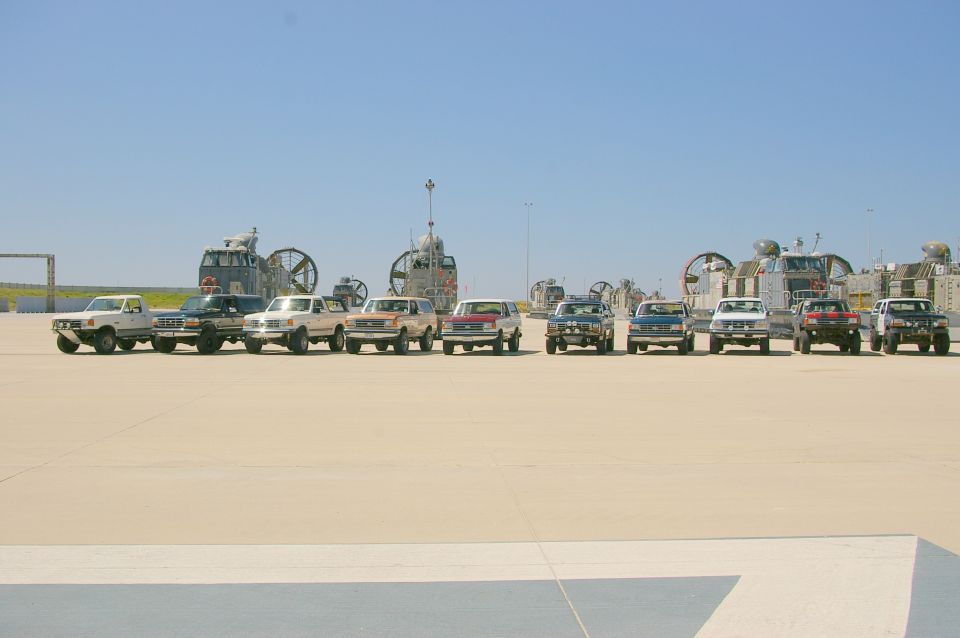 Line of 1986 Ford Bronco SUVs and Ford F-Series trucks parked on pavement with hovercraft in background