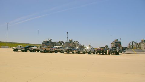 1986 Ford Bronco - Several SUVs are parked in a line on a paved surface, with large hovercraft visible behind them under a clear blue sky.